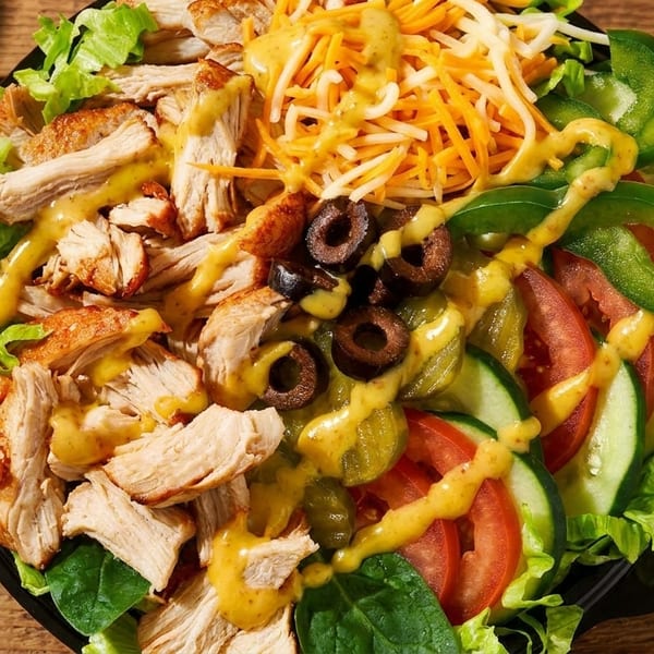 Close-up of a vibrant salad with grilled chicken, shredded cheese, olives, and vegetables, drizzled with dressing.