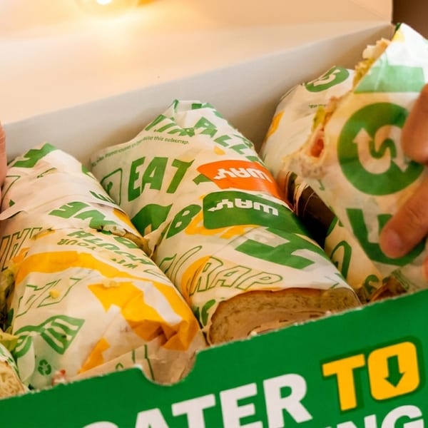 Hands reaching into a Subway catering box filled with individually wrapped sandwiches.
