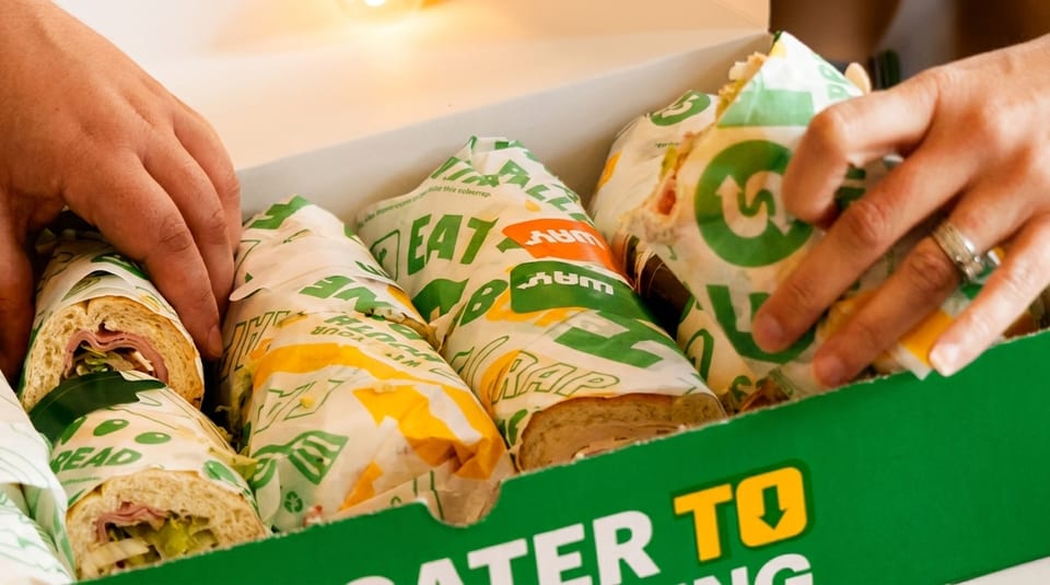 Hands reaching into a Subway catering box filled with individually wrapped sandwiches.