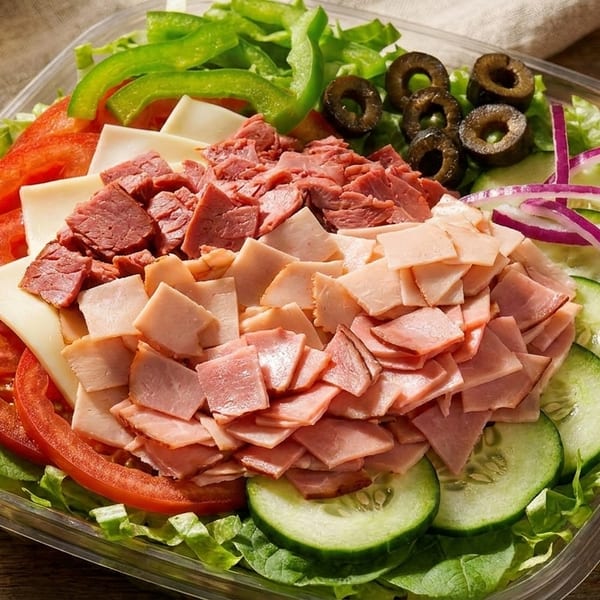 A clear plastic container filled with a fresh salad, featuring various meats, cheeses, and vegetables.