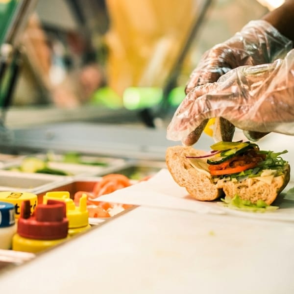 Gloved hands preparing a Subway sandwich with fresh vegetables and condiments in the foreground.