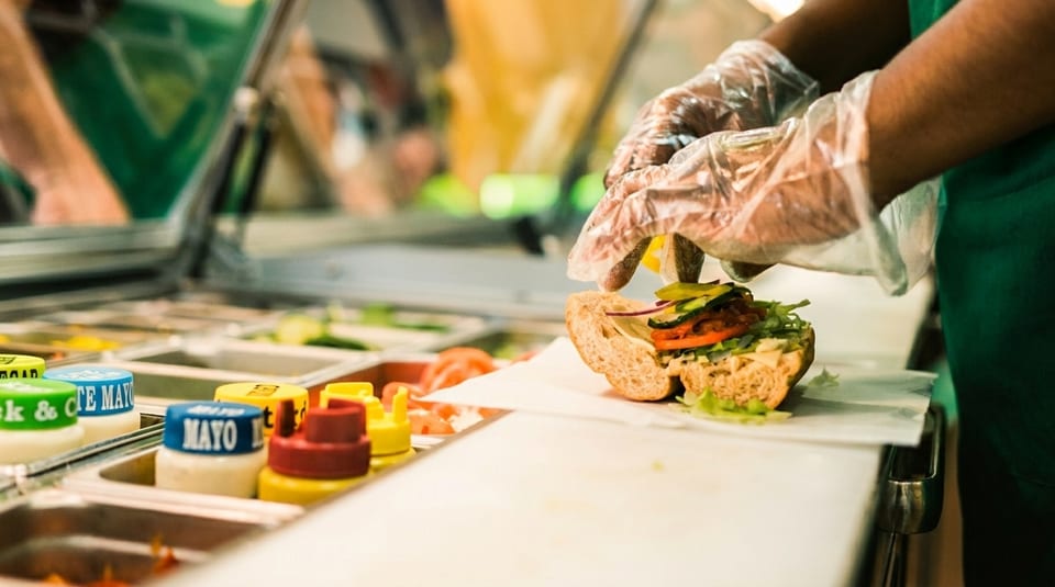 Gloved hands preparing a Subway sandwich with fresh vegetables and condiments in the foreground.