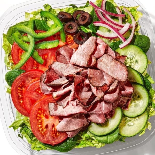 A vibrant salad in a clear plastic container, featuring sliced roast beef, tomatoes, cucumbers, and olives.