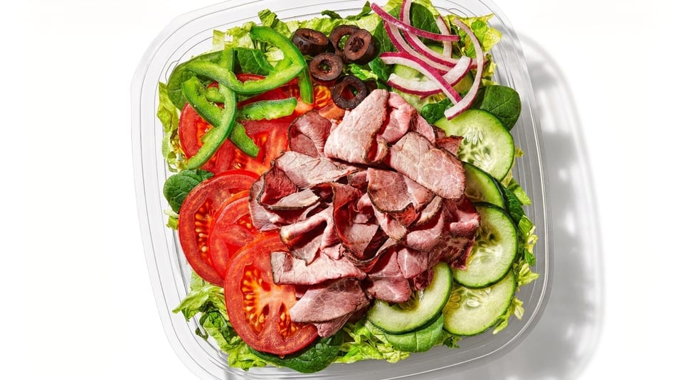 A vibrant salad with sliced roast beef, tomatoes, cucumbers, olives, peppers, and red onions in a clear container.