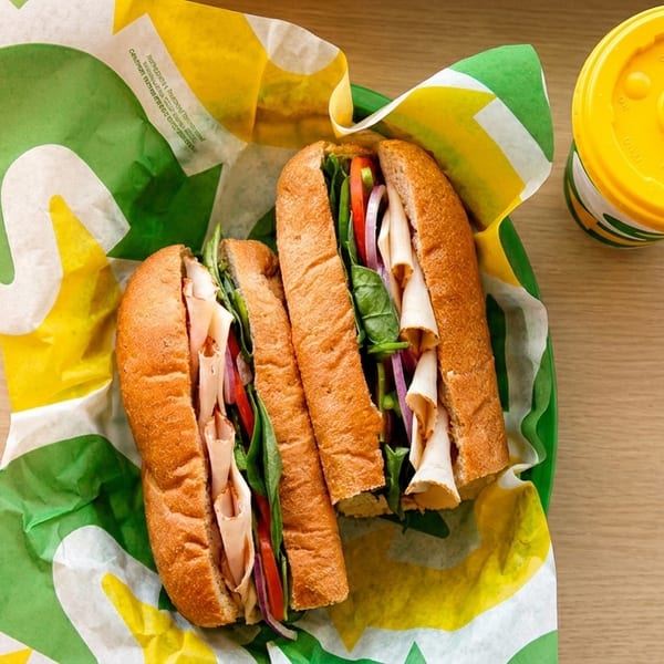 Two Subway sandwiches with turkey, vegetables, and a drink on a wooden table.