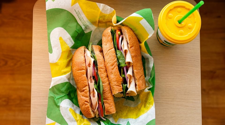 Two Subway sandwiches with turkey, vegetables, and a drink on a wooden table.