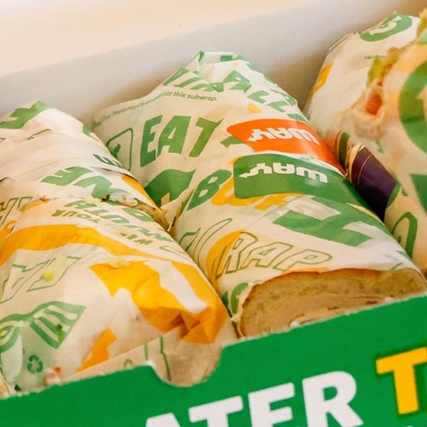 Hands reaching into a green Subway catering box filled with individually wrapped submarine sandwiches.