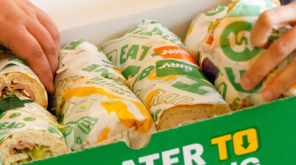 Hands reaching into a green Subway catering box filled with individually wrapped submarine sandwiches.